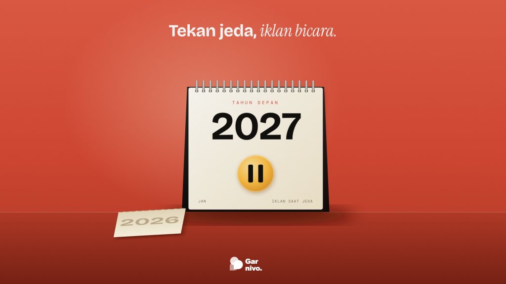 Kalender meja spiral menampilkan 2027 dengan lencana jeda amber di dinding merah, lembar 2026 yang disobek tergeletak di meja.