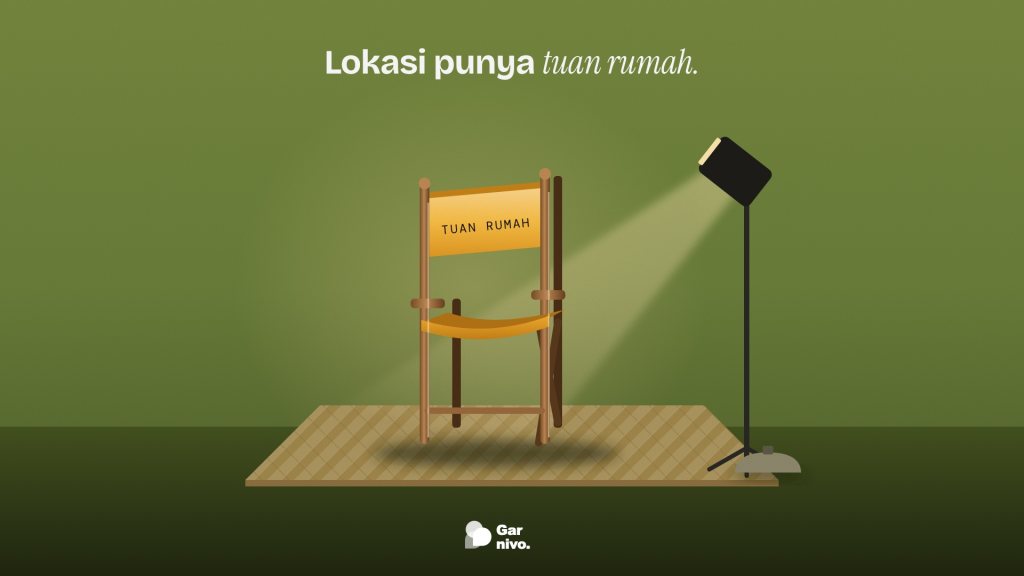 Kursi sutradara kayu dengan kain sandaran amber bertuliskan tuan rumah berdiri di atas tikar anyaman, disorot lampu studio di latar hijau lumut.
